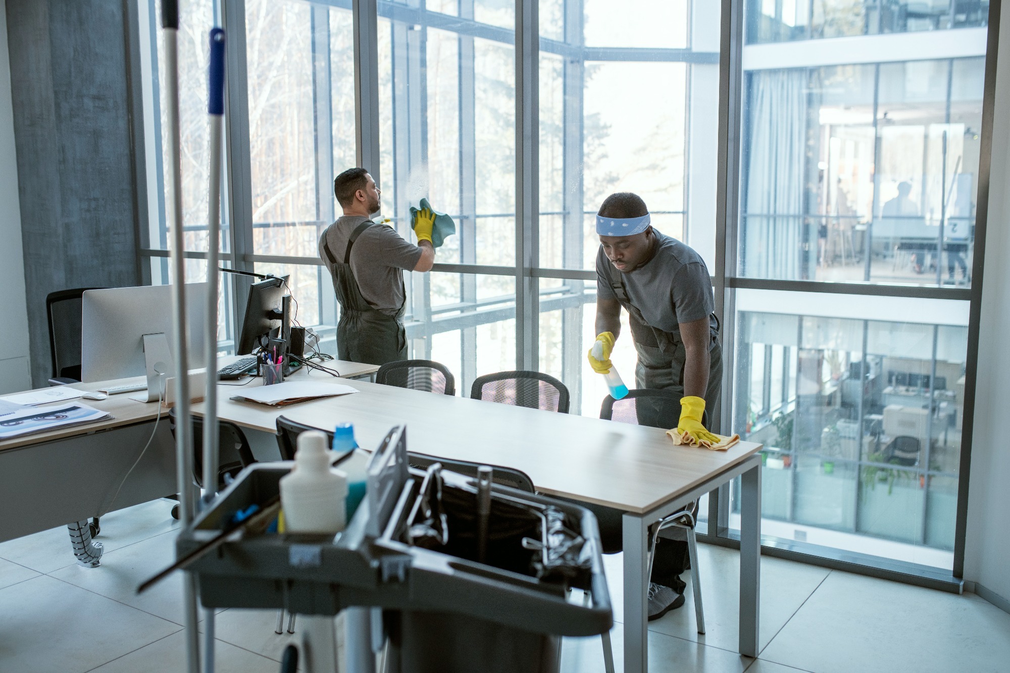 A team of professional cleaners wearing uniforms and gloves, diligently working to clean and sanitize a spacious office environment, focusing on desks, electronic equipment, and flooring.