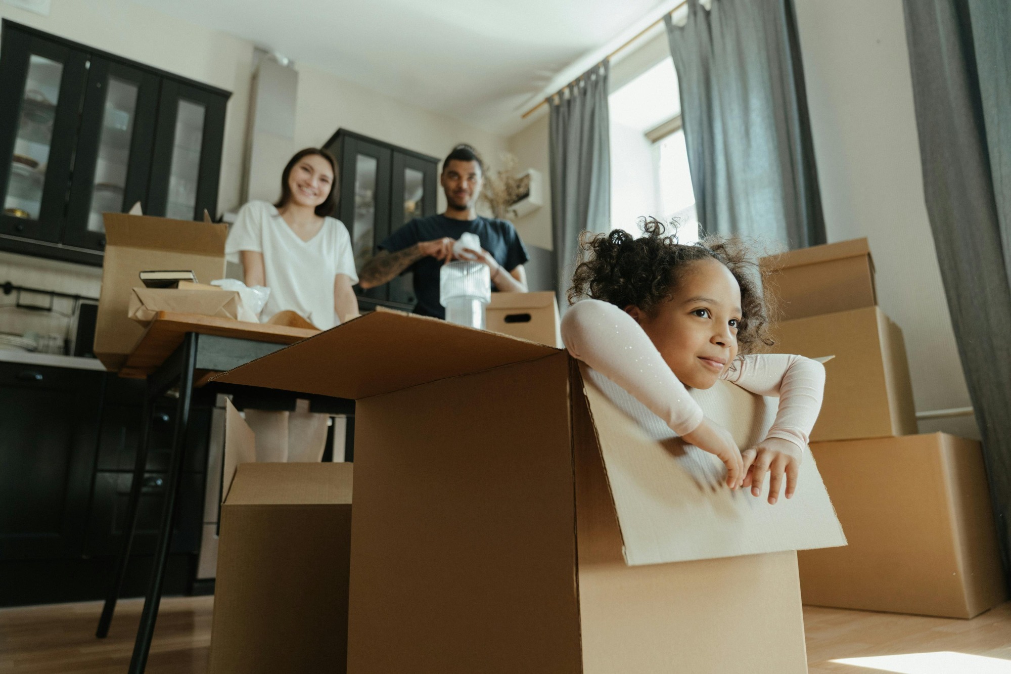 A smiling family of three, with two children, standing in front of a moving truck outside their new home in Rhode Island, surrounded by boxes.