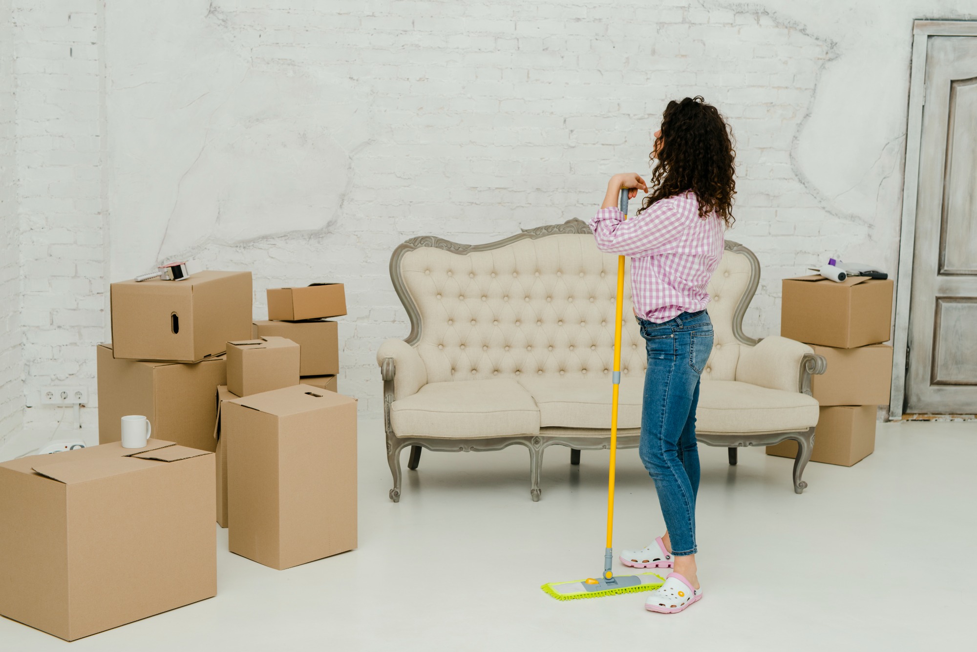 A woman stands confidently in a sunlit room, holding a mop, with moving boxes scattered around her, symbolizing the beginning of a new chapter in her Rhode Island home