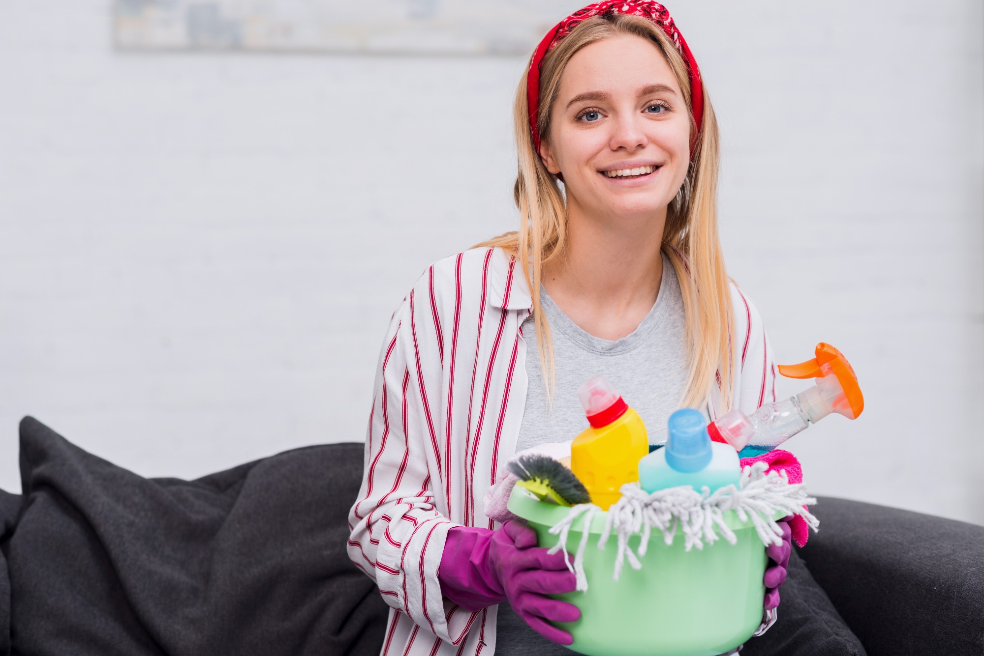 A smiling maid holding a bucket filled with cleaning supplies including sprays, sponges, and cloths